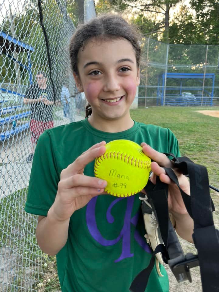 Person holding a bright yellow softball on a field, smiling near a chain-link fence.
