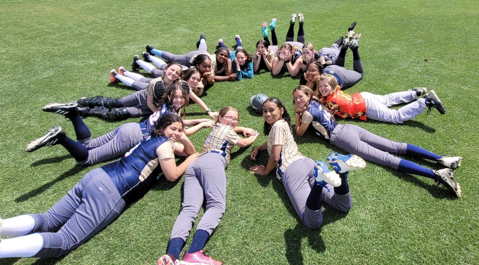 Group of people lying in a circle on grass, reaching hands together and smiling outdoors.