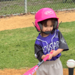 Tee-ball player in a pink helmet holding a pink bat, standing on a baseball field near a tee.