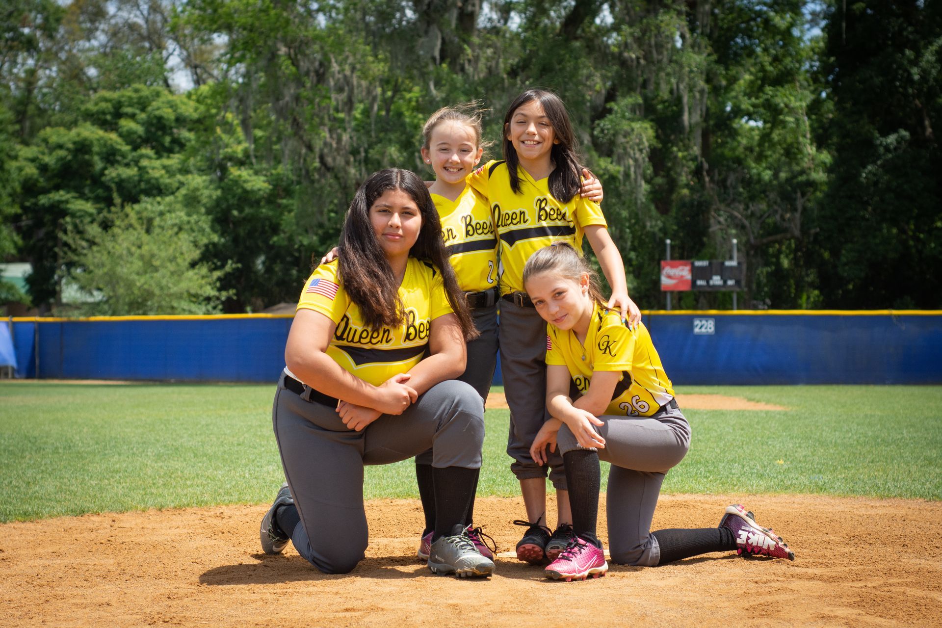 Three softball players in yellow jerseys pose on a field, one kneeling and two standing behind her.