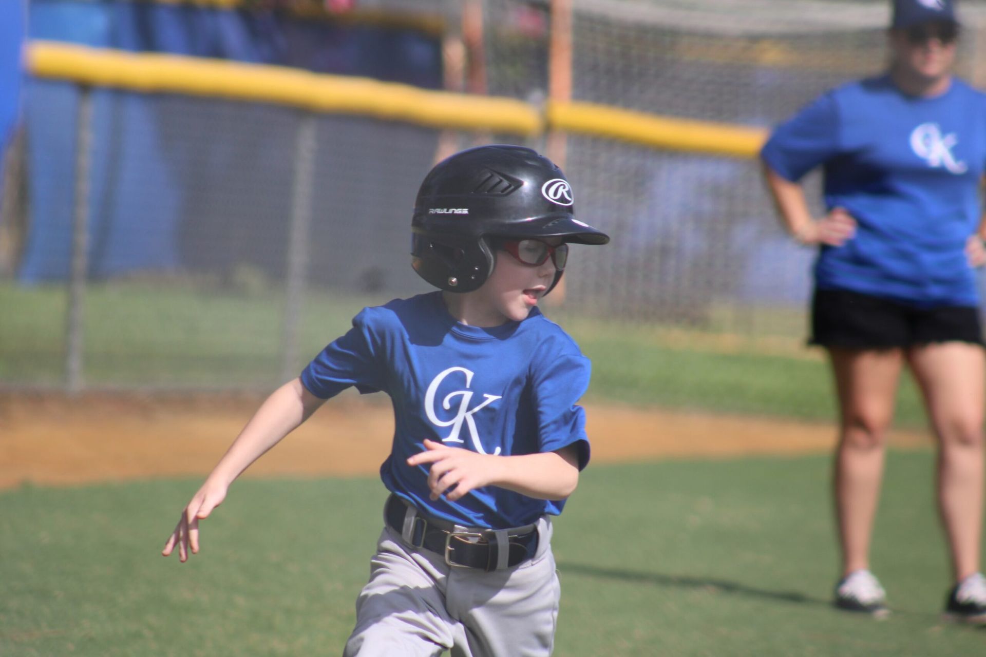 Child in a blue shirt and helmet runs on a baseball field, with a coach in the background.
