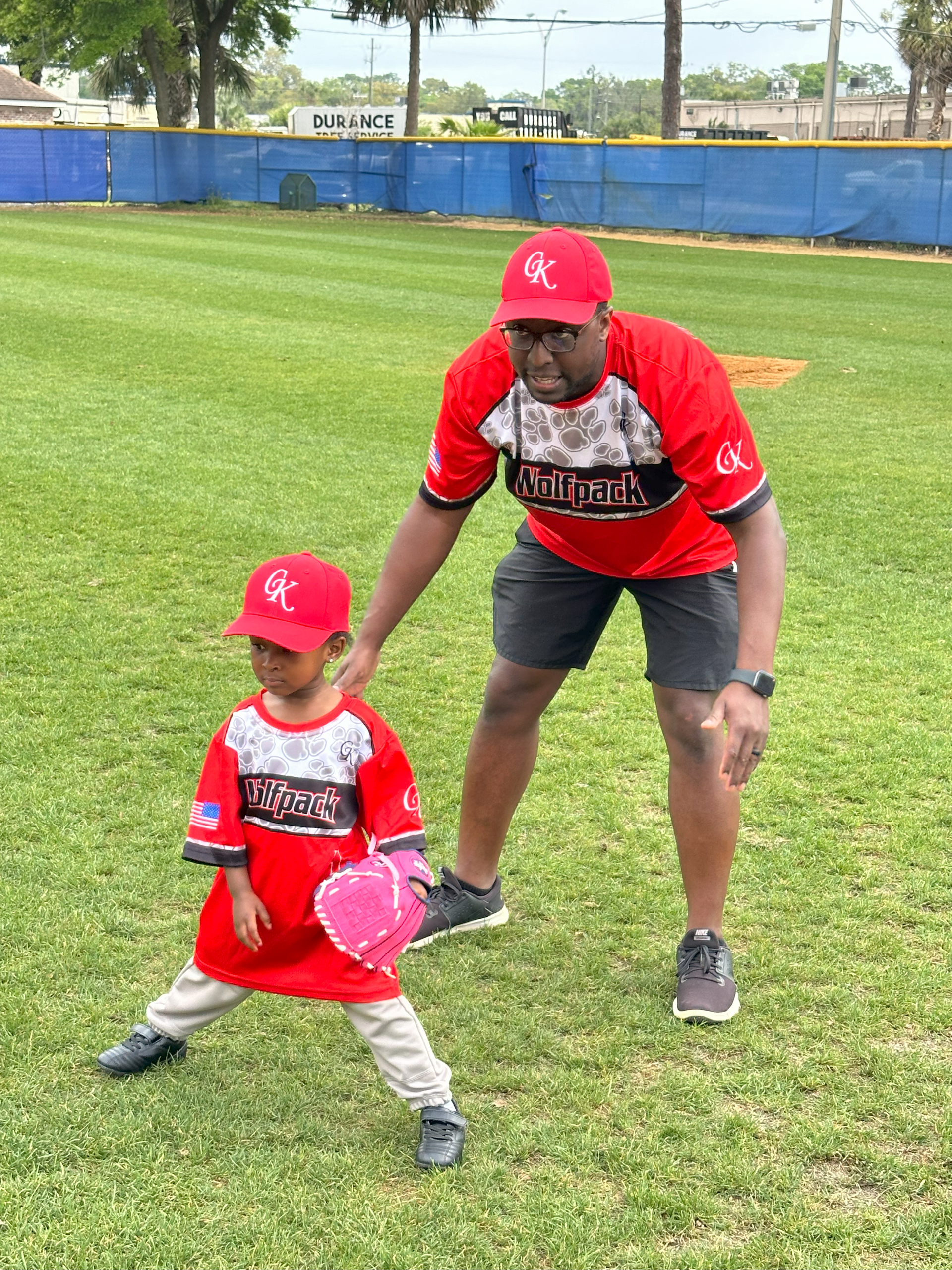 Coach and a tee-ball player in red uniforms posing on a grassy field, one child holding a pink glove