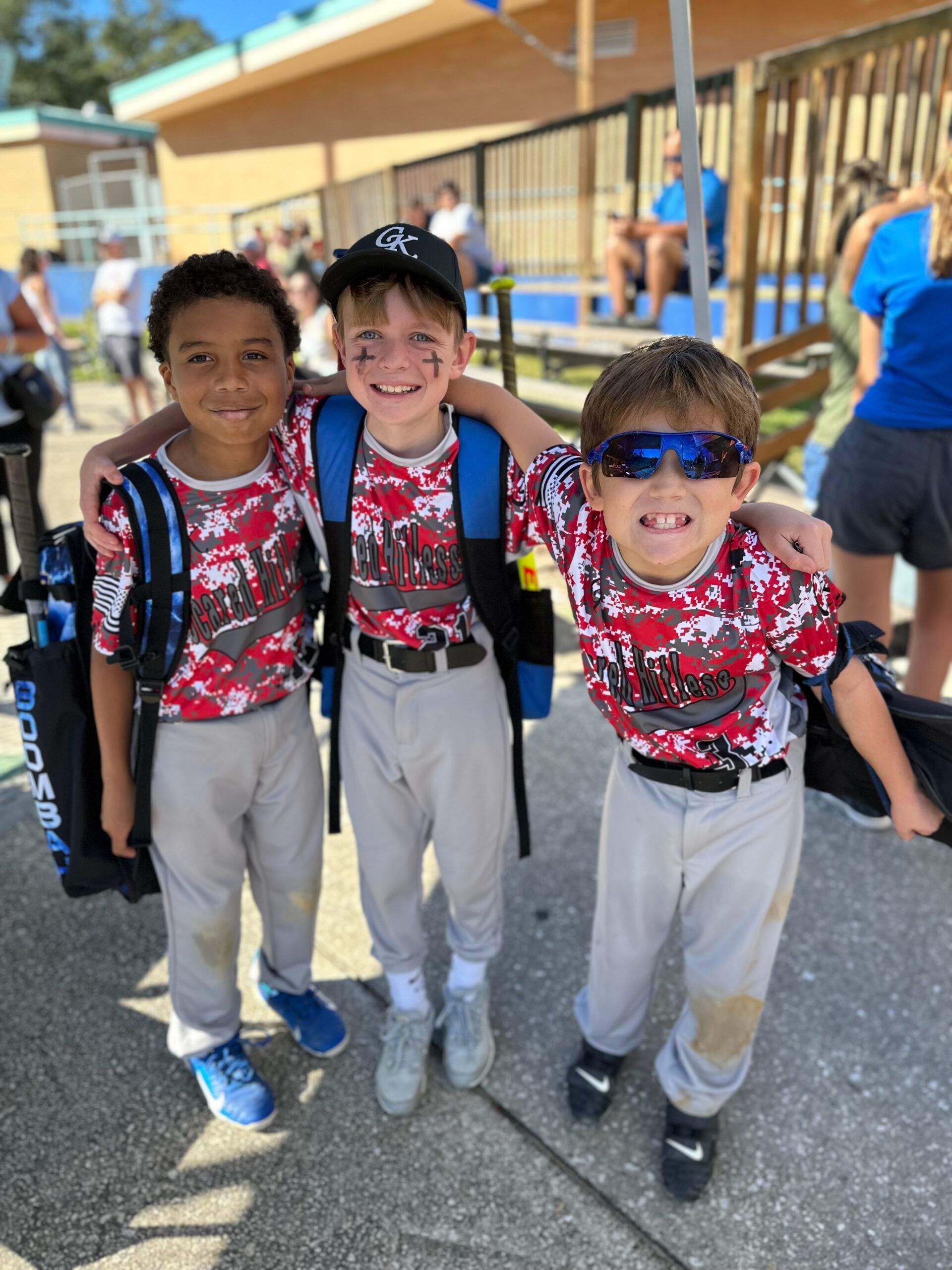 Three children in matching red jerseys and gray pants stand smiling outdoors by a school playground.