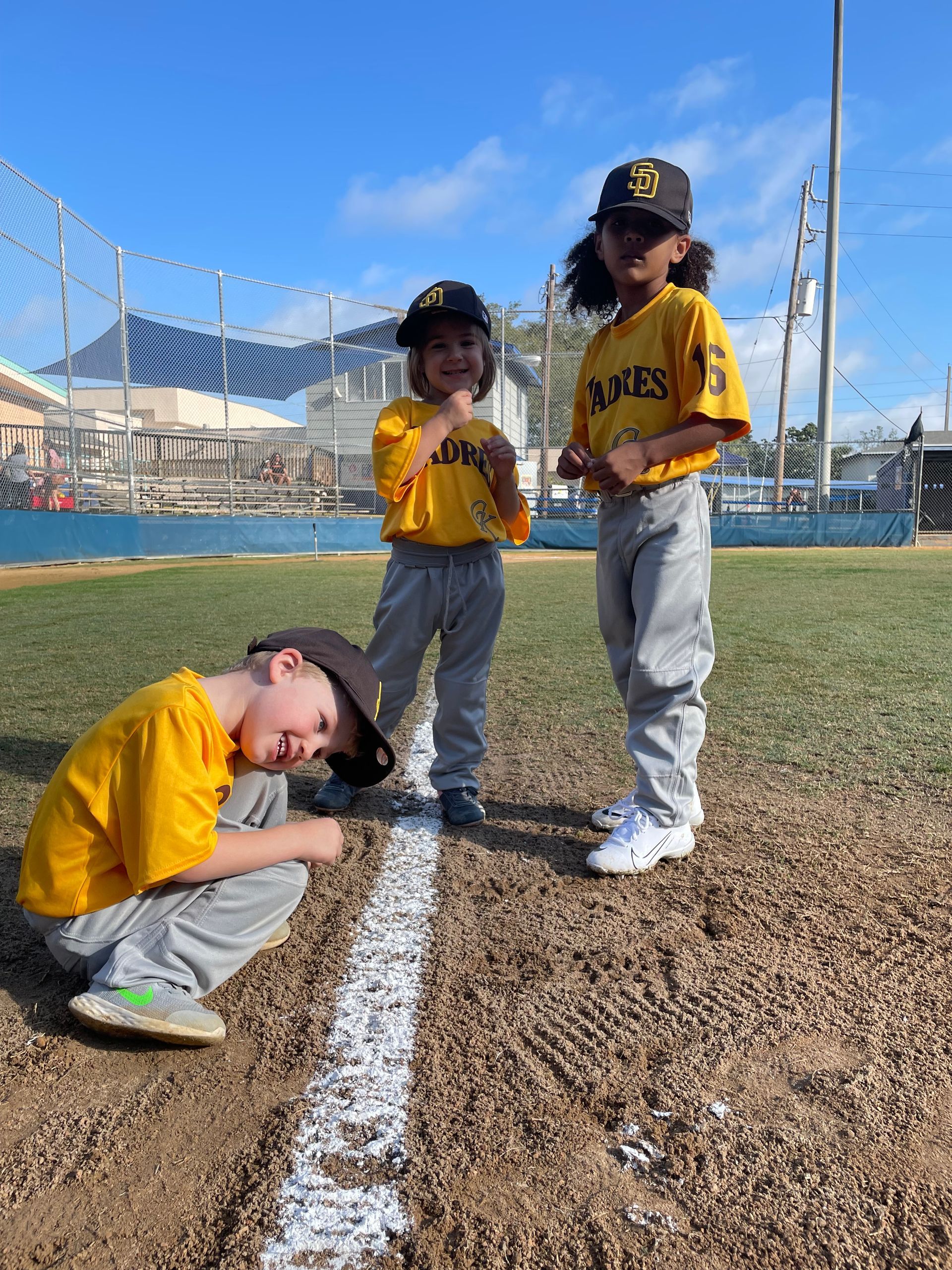 Three children in yellow sports uniforms on a gravel field by a marina, one crouching near a white line.
