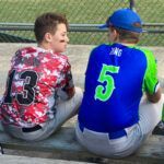 Two people in sports jerseys sit on a bench, talking outdoors near a fence.