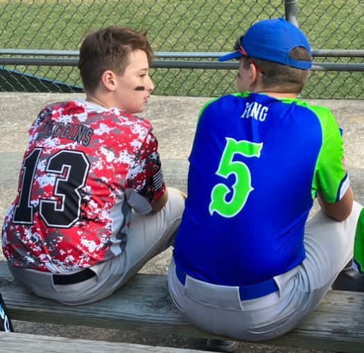 Two baseball players in red and blue jerseys sit on a bench near a chain-link fence.