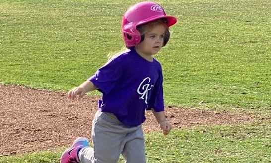 Child in a pink helmet and purple shirt kneels on grass beside a dirt baseball field.