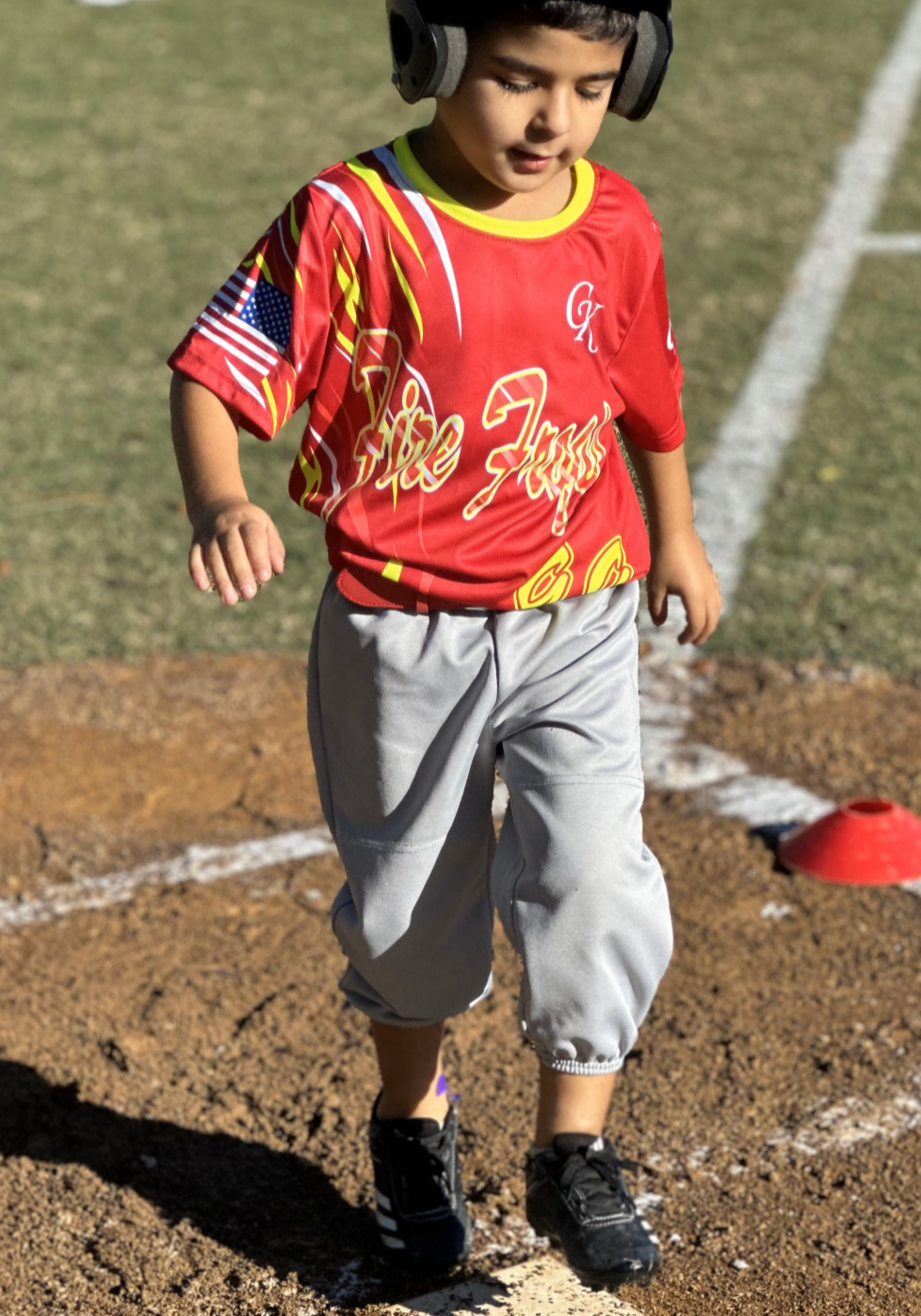 Tee-ball player wearing a red jersey and gray pants walking on a dirt sports field with headphones on