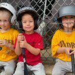 Three Tee-ball players in baseball helmets and T-shirts standing with bats in front of a chain-link fence