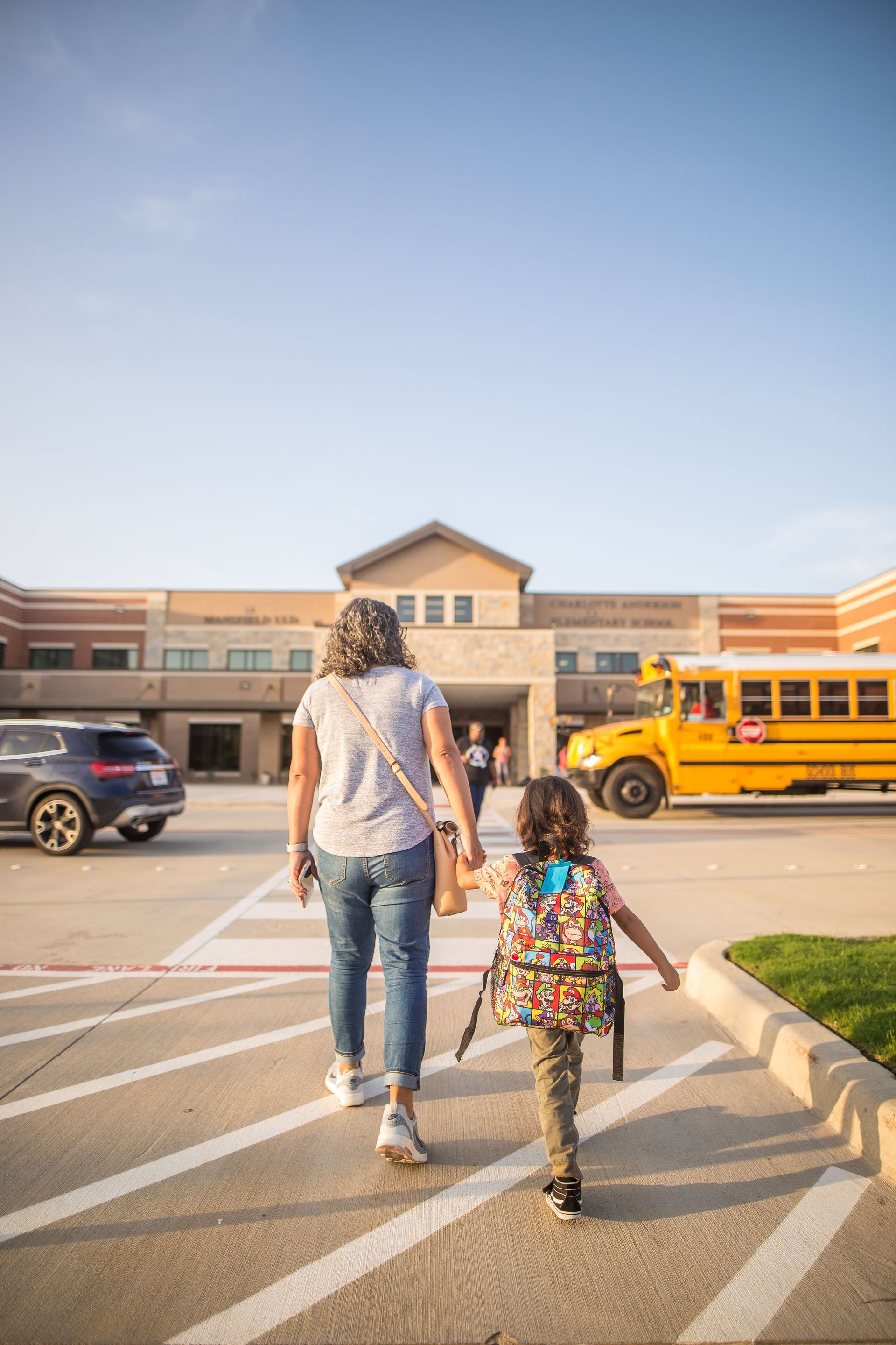 Mom walking child through school parking lot. Yellow school bus parked outside school.