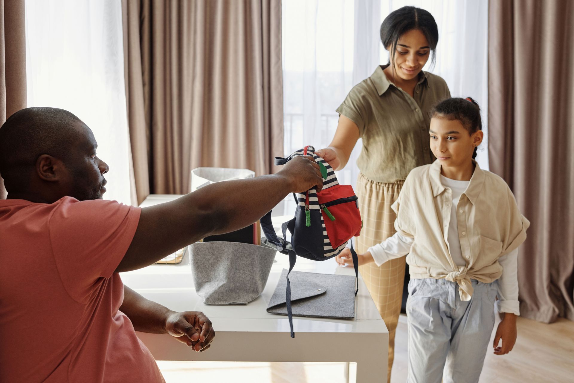 Man in orange shirt handing backpack with red and white stripes to little girl and mother