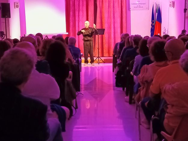 Un oratore su un piccolo palco si rivolge a un pubblico in un auditorium illuminato di rosa.