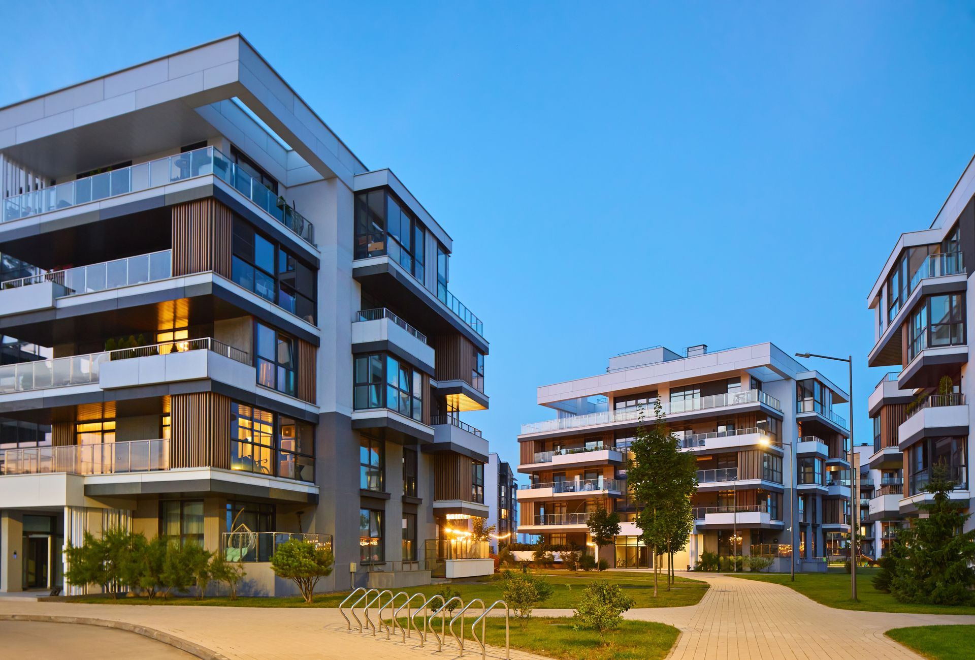Modern apartment buildings at dusk with lit windows, balconies, and a courtyard path under a blue sky