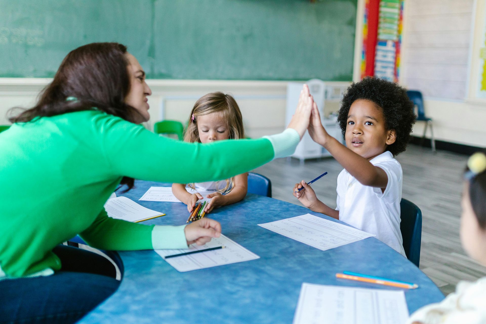 Teacher and students high-five at a classroom table with papers and pencils.