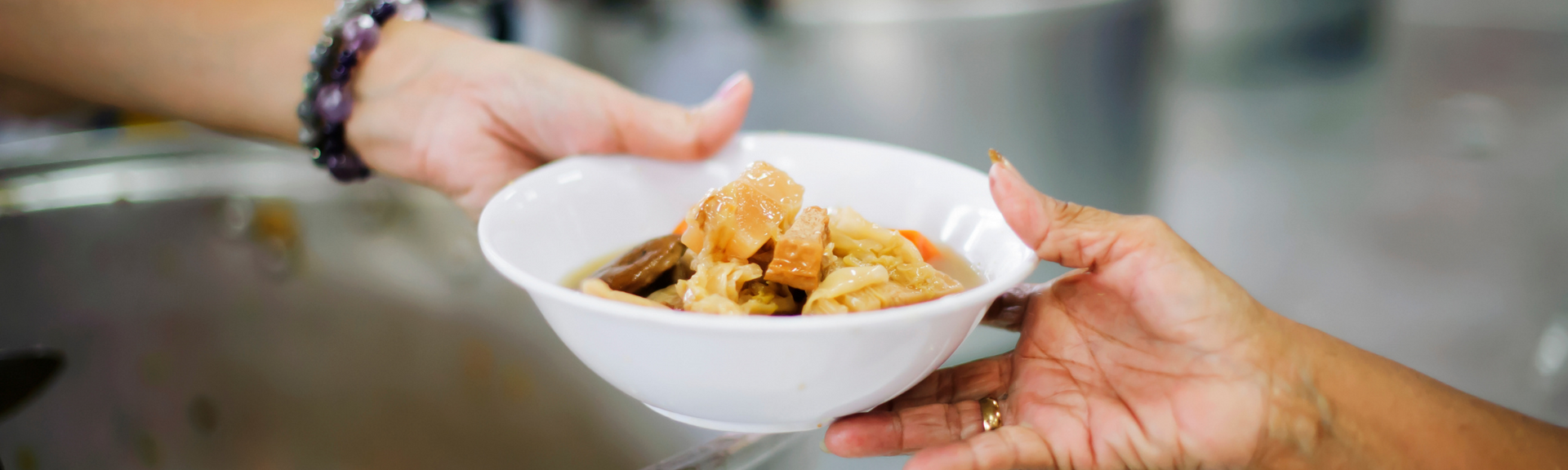 Hands passing a white bowl of pasta in a blurred kitchen setting