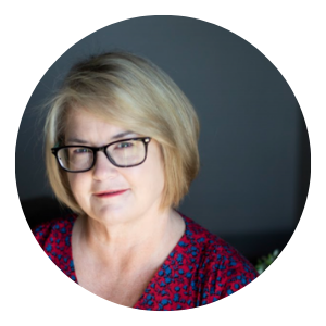 Portrait of a person with glasses and short blonde hair, wearing a red patterned top against a dark background