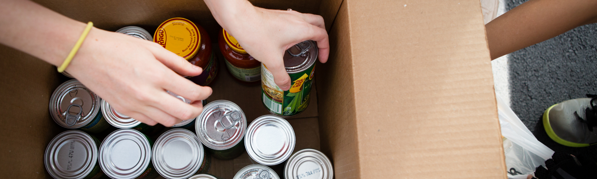 Hands sorting canned food in a cardboard box, with several other cans visible nearby.
