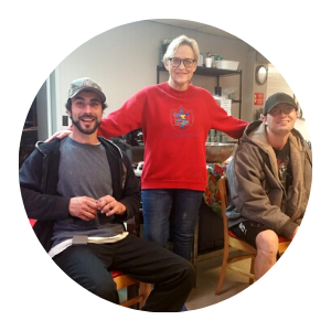 Three people posing indoors, with the center person in a red sweater standing between two seated people.
