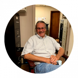 Smiling man seated in an office chair, wearing a light striped shirt in a hallway or office setting