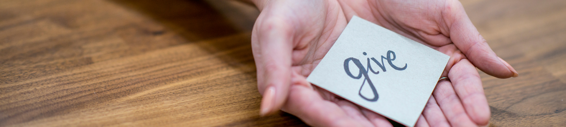 Hands holding a white card with the word “give” on a wooden table.