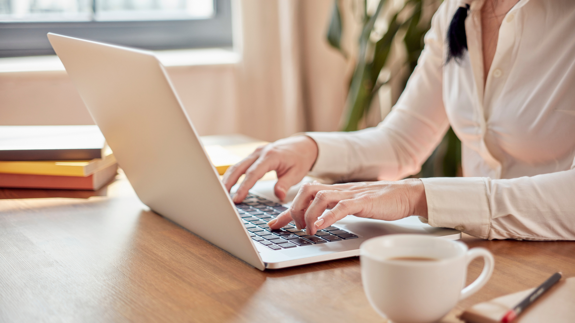 A woman is typing on a laptop computer while sitting at a desk with a cup of coffee.