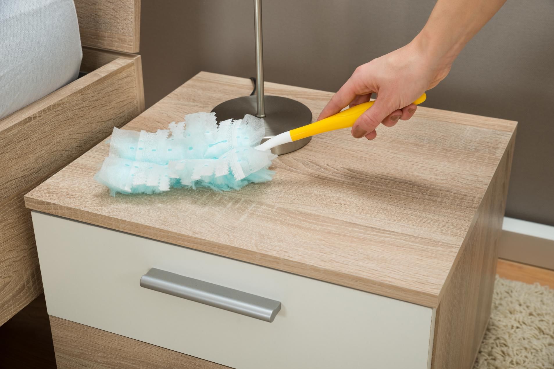 A hand uses a yellow-handled duster with light blue fibers to clean a light wood-grain bedside table.