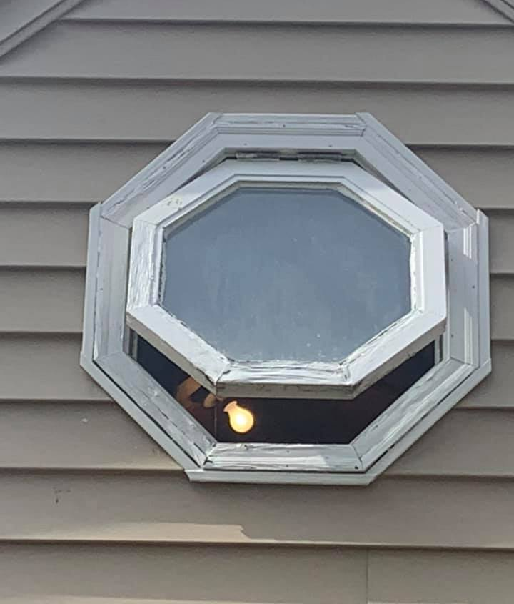 Octagonal window on a tan house. The inner pane is open, revealing a lightbulb. White trim shows wear.