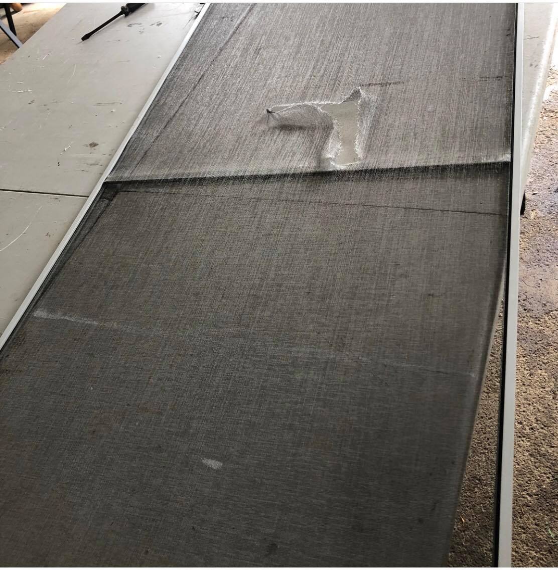 A damaged window screen laid on a white table. A hole is visible on the upper part.