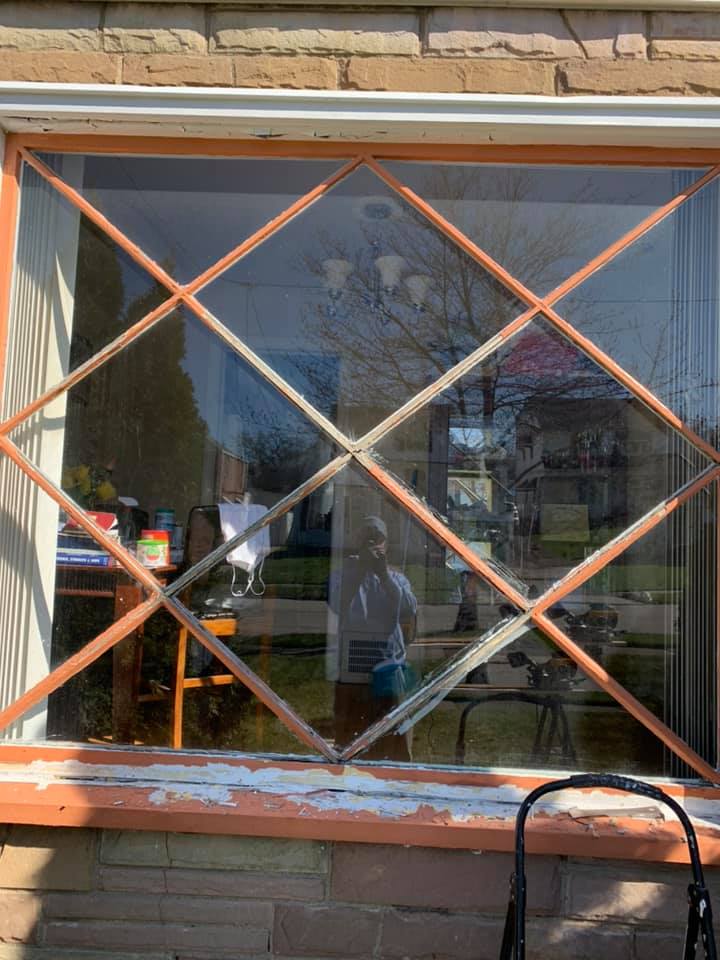 Window with diamond-shaped panes, peeling orange paint on the frame, outdoor view with a person reflected in the glass.