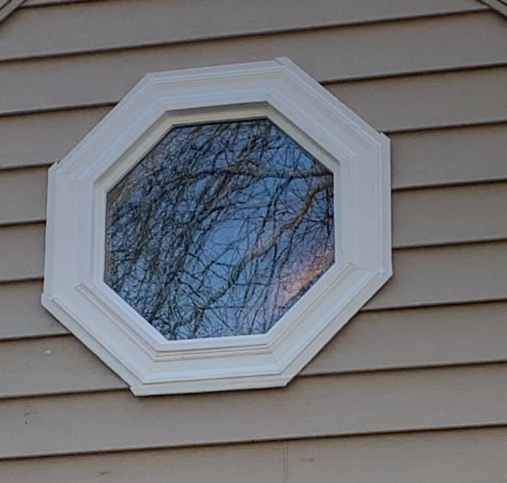 Octagonal white-framed window on tan siding reflecting tree branches against a blue sky.