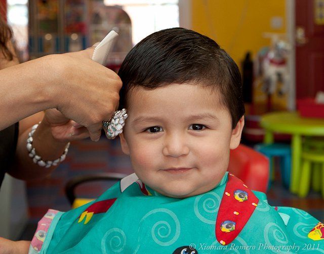 Child getting a haircut in a salon, wearing a turquoise cape and smiling at the camera.