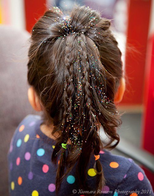 Child seen from behind with a sparkly ponytail, wearing a dark polka-dot shirt, indoors.