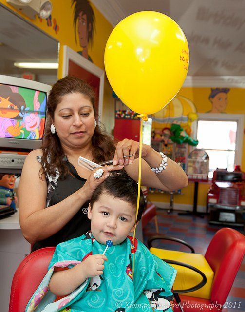 Hairdresser cutting a child's hair with a yellow balloon nearby in a bright salon
