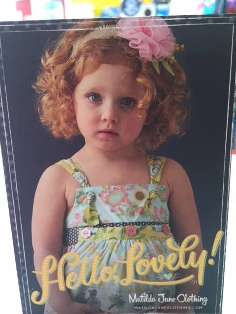 Young child with curly hair and pink flower headband, wearing a patterned dress against a dark background.