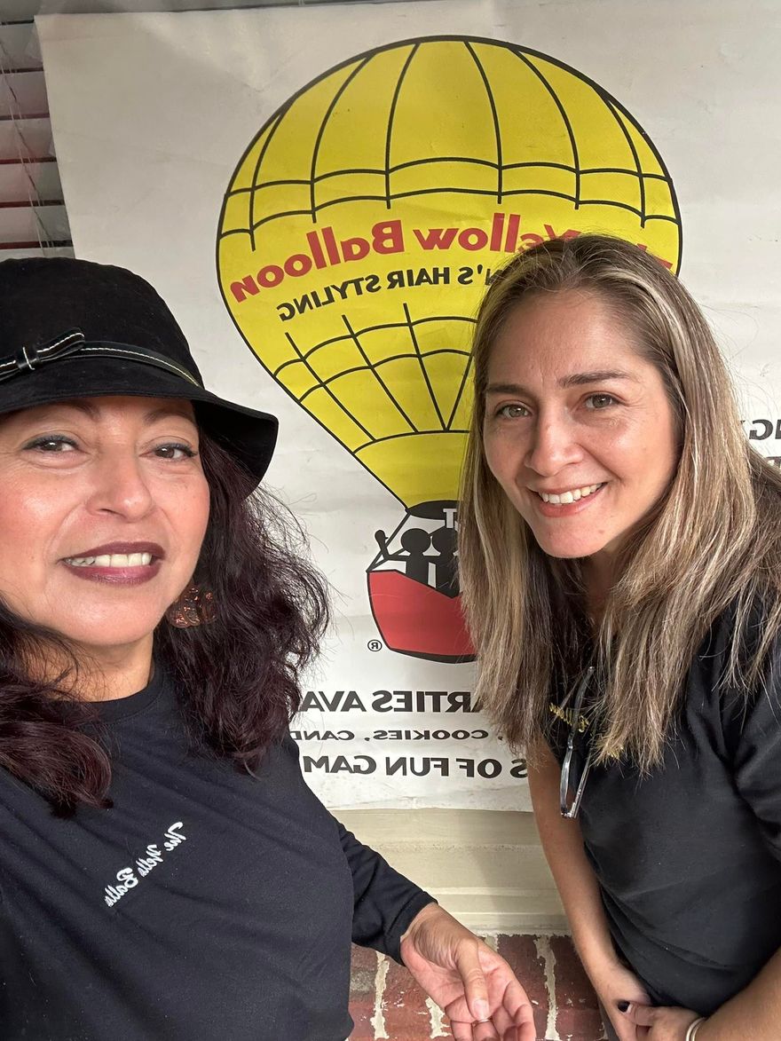 Two women smiling in black shirts posing in front of a yellow hot air balloon sign.