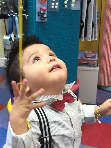 Child with dark hair in a ponytail, seen from behind, wearing a polka-dot shirt indoors.