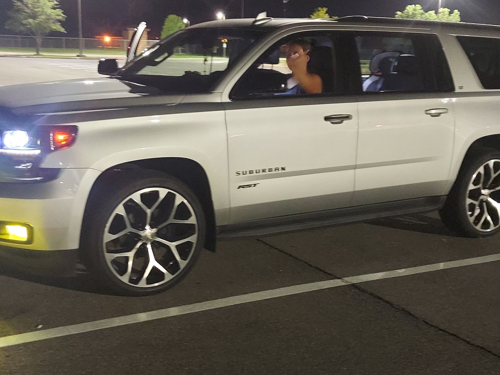 White SUV with headlights on parked at night, driver visible through the open window.