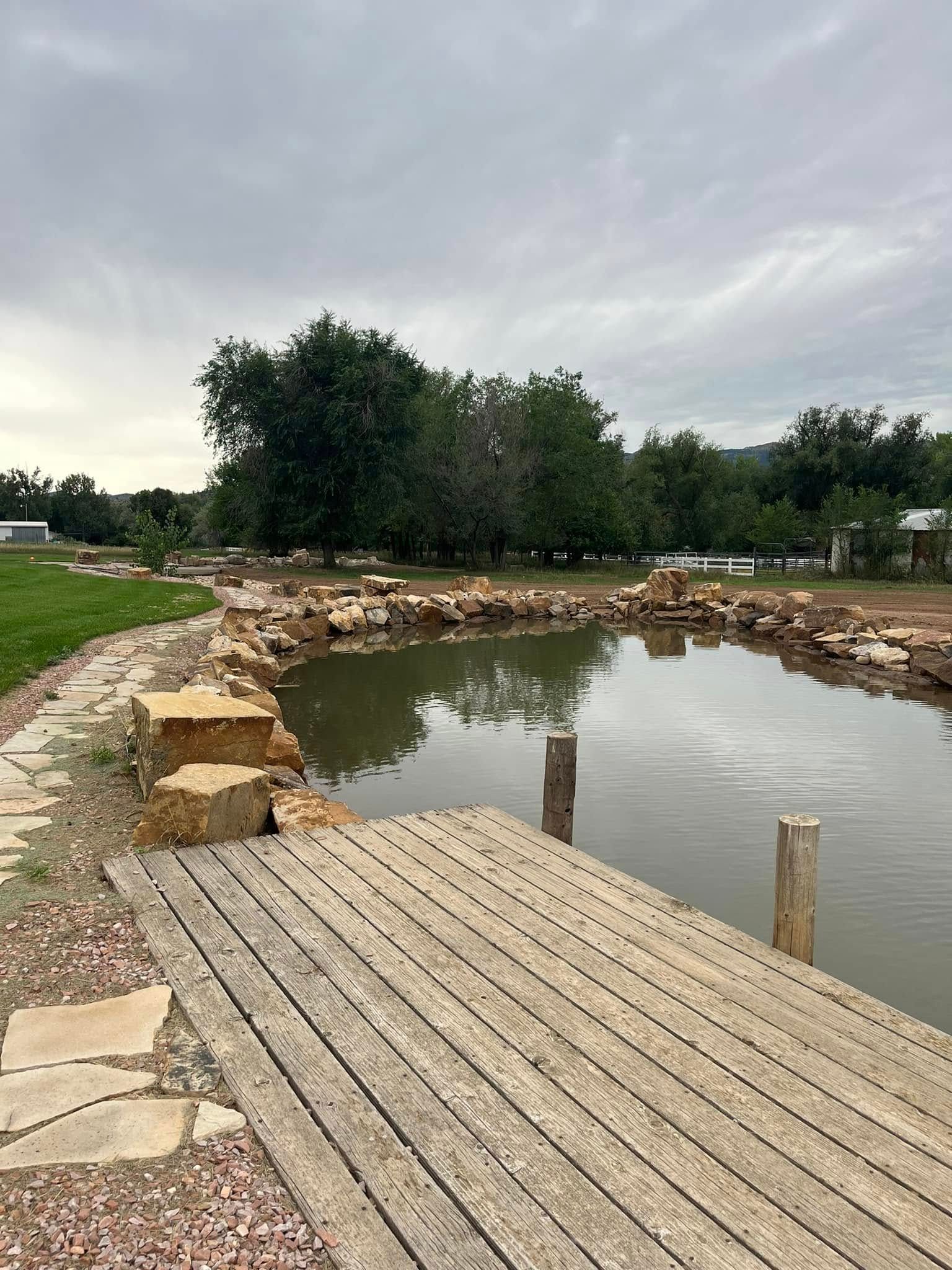 There is a wooden dock next to a pond with trees in the background.