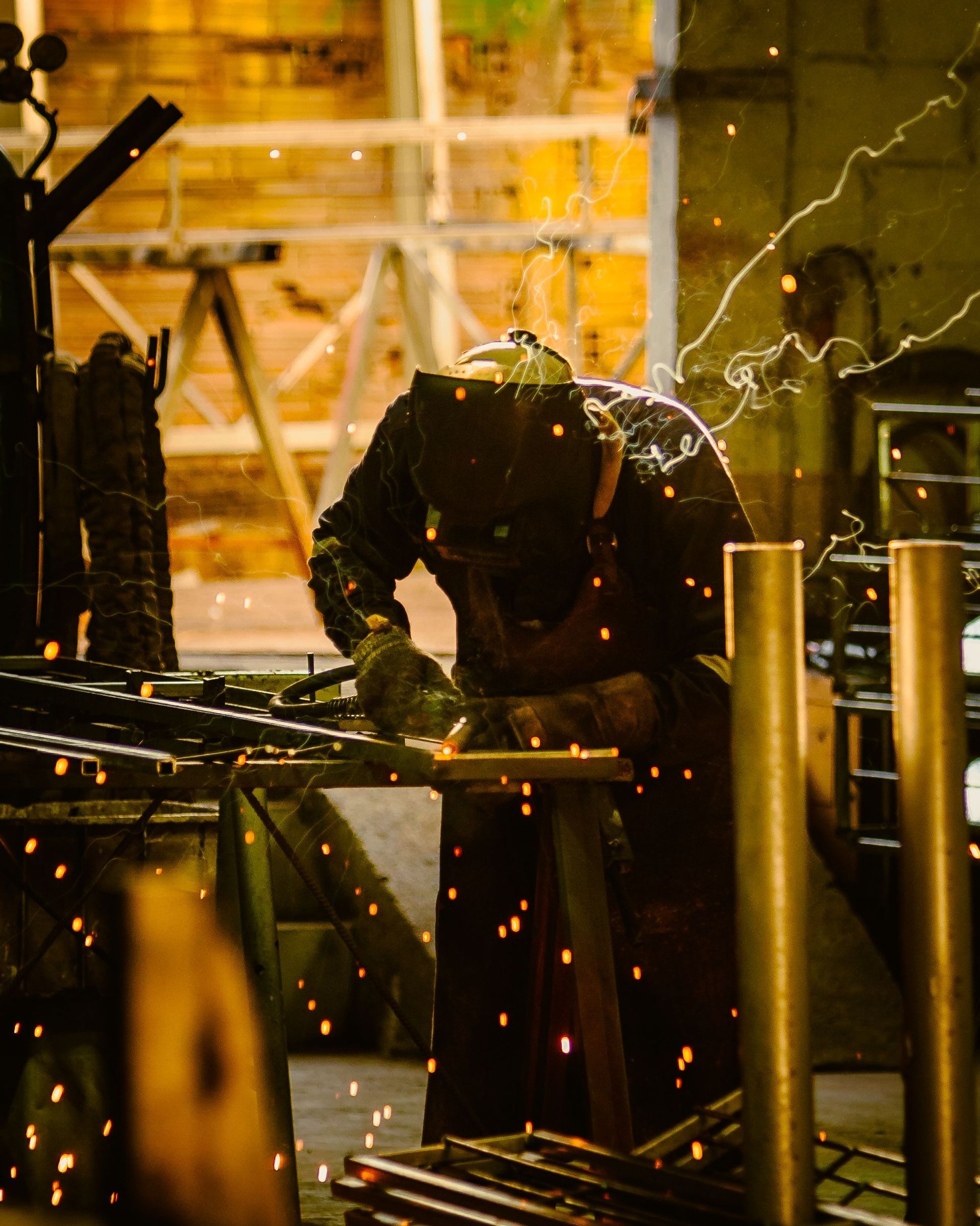 Welder in a dim workshop sparks metal on a workbench, with orange sparks flying around.