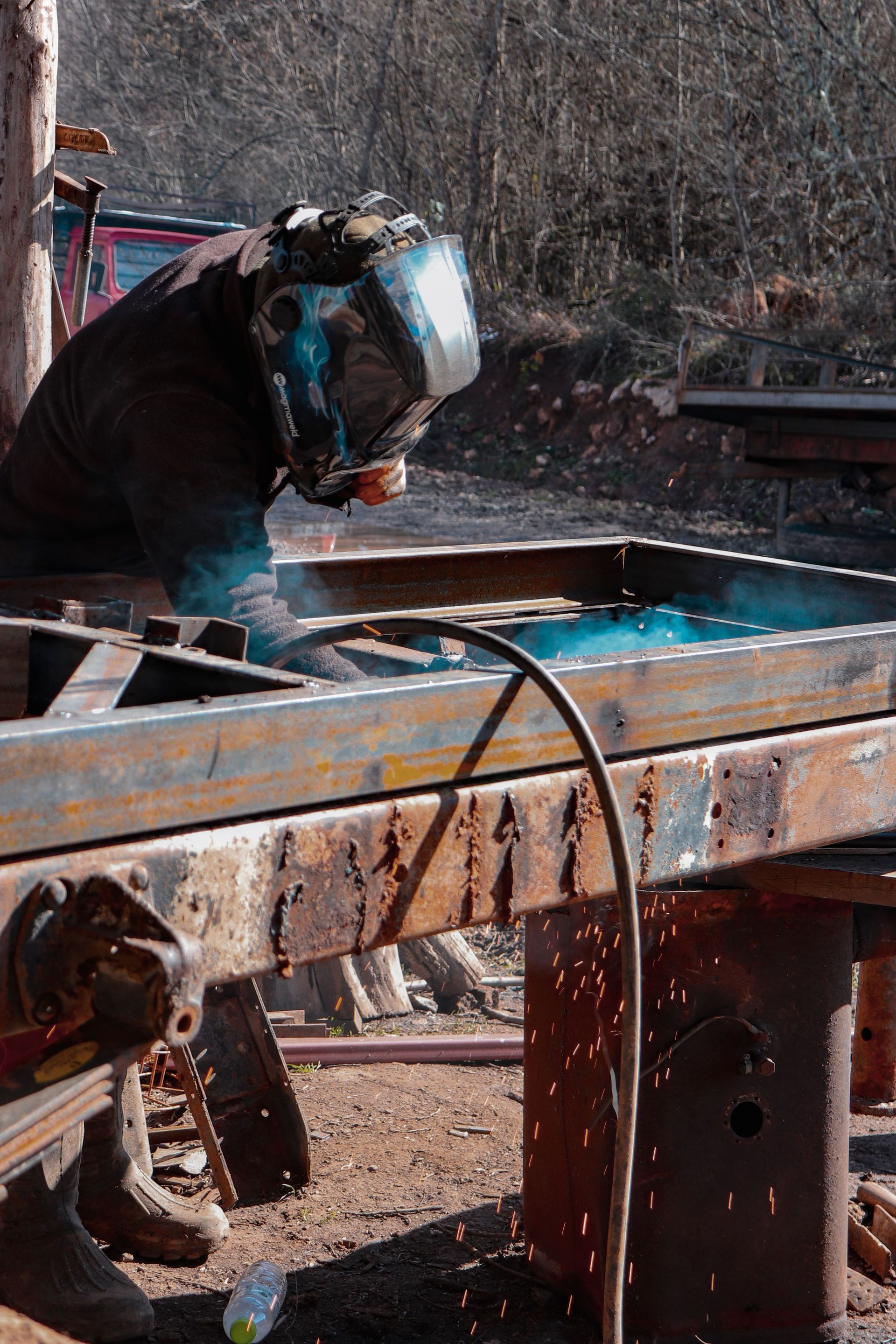Person welding at a rusty metal table outdoors, with bright sparks and a face shield.