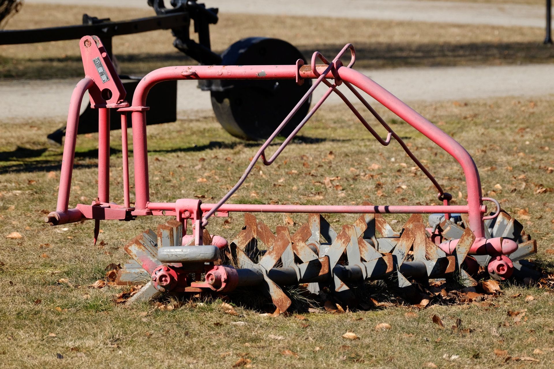 Pink tractor-mounted tiller on grass, with exposed metal blades and a curved frame.