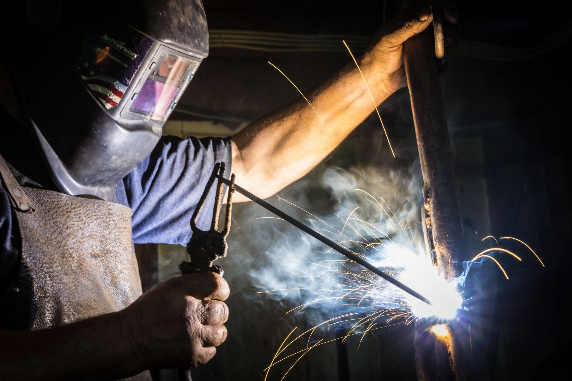 Welder in protective mask welding metal, bright sparks and blue arc in a dark workshop