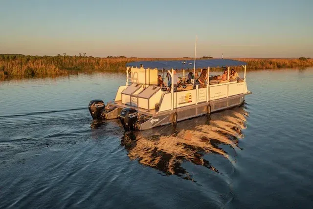 Pontoon boat cruising across calm water at sunset, with trees lining the shore.