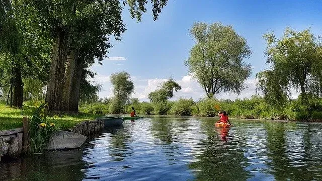 Calm river with two people paddling, trees lining the banks, and bright blue sky above