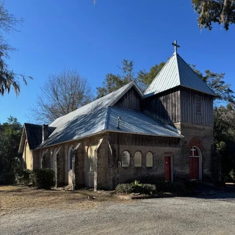Small rustic church with a metal roof and cross, set beside a gravel lot under a clear blue sky