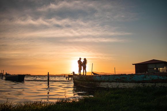 Two people stand on a dock at sunset, silhouetted against orange sky over calm water.