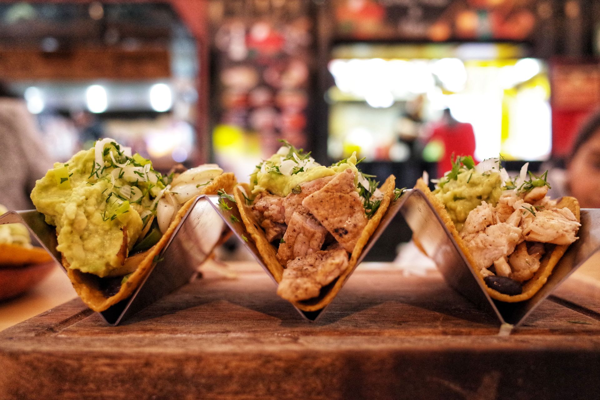 Three loaded tacos in paper holders on a wooden table, with a blurred restaurant background