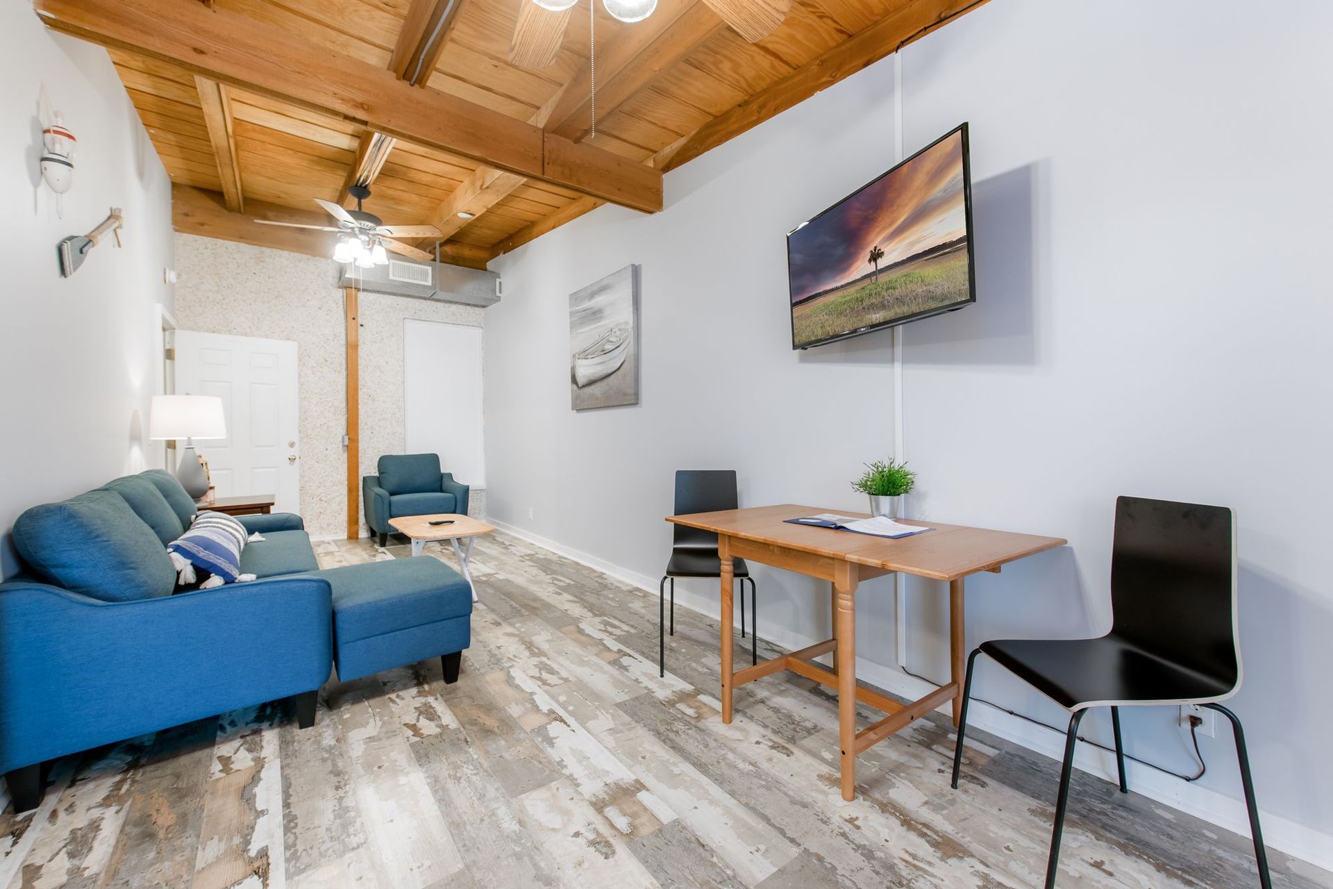 Modern living room with blue sofa, dining table, wall TV, and wood ceiling with skylight.