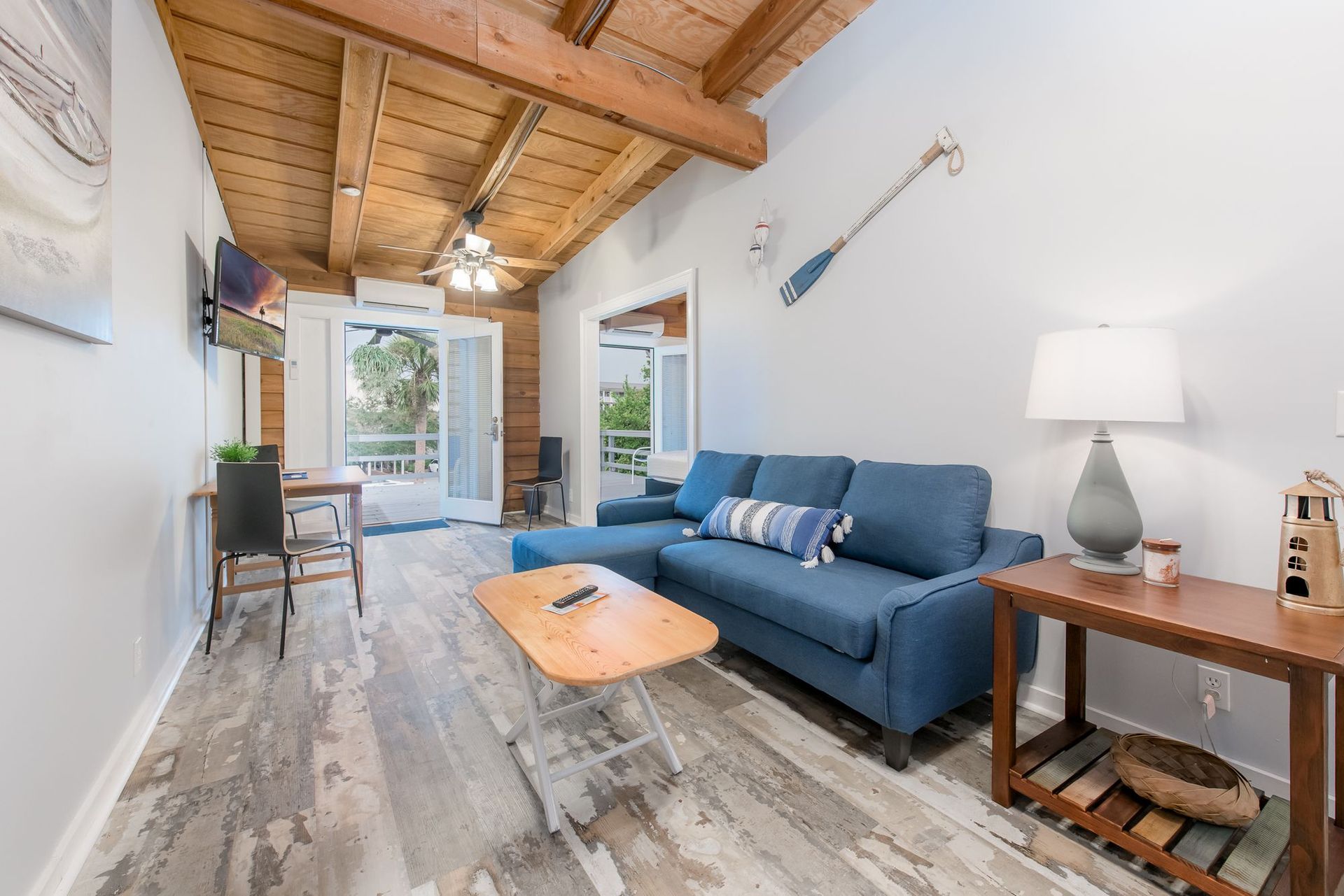 Bright living room with blue sofa, wooden coffee table, side lamp, and vaulted wood ceiling, opening to a patio