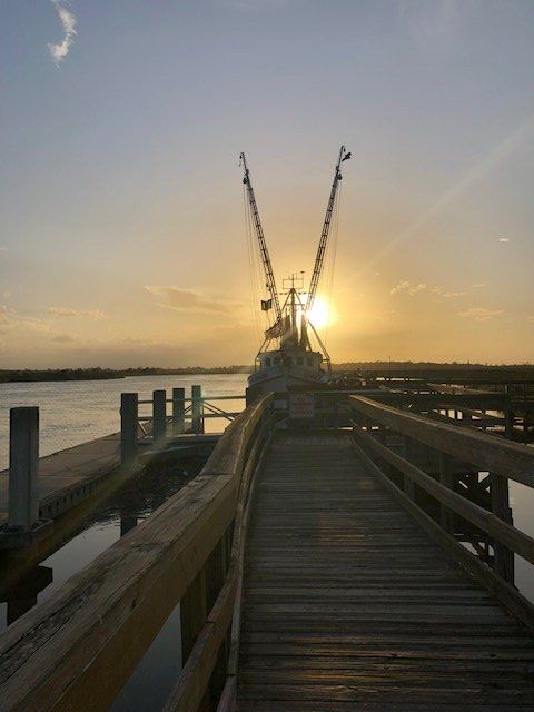 Wooden dock at sunset with fishing boats and calm water.